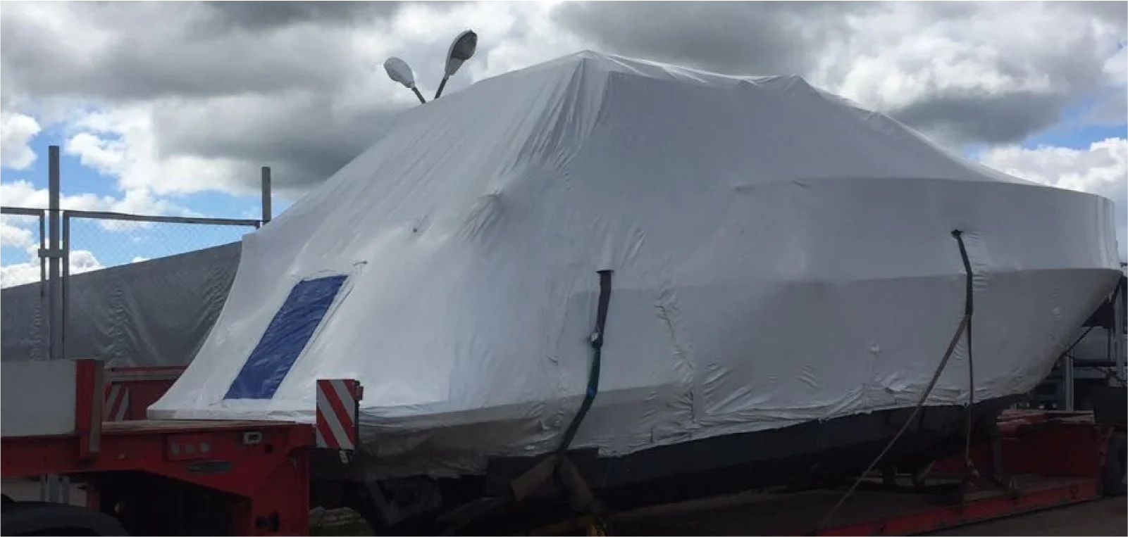A large motor yacht securely wrapped in white protective film, loaded onto a red low-loader trailer for oversized cargo transport from Italy to Norway by Lazarus.